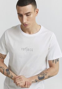 Hvid bomuld t-shirt med ordet "reflect" i grå tekst, rund hals, korte ærmer. Individ med tatoveringer holder hænderne sammen.