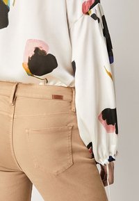 Blouse blanche avec des motifs abstraits colorés, associée à un pantalon beige ajusté avec une poche arrière et une petite étiquette en tissu près de la taille.