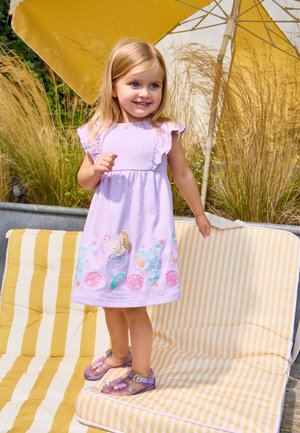 Fiatal lány lila ruhában, sellő hímzéssel, sárga-fehér csíkos párnákon áll egy sárga esernyő alatt, mögötte magas fűvel.