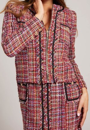 Woman wearing red and pink plaid tweed jacket and skirt with black trim and gold buttons, hand near face and resting on hip.