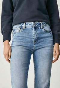Jasnoniebieskie dżinsy o dopasowanym kroju, z pięcioma kieszeniami, z zapięciem na guzik i wyblakłą fakturą w okolicach ud, zestawione z ciemnym swetrem.