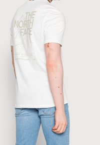 Muž v bílém tričku The North Face s grafikou hory a nápisem "Nikdy nepřestávejte objevovat", v kombinaci se světle modrými džíny.