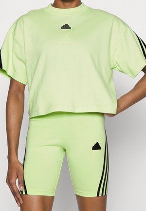 Persoon draagt lichtgroen cropped T-shirt en bijpassende strakke korte broek met zwarte Adidas-logo's en strepen op mouwen en benen.