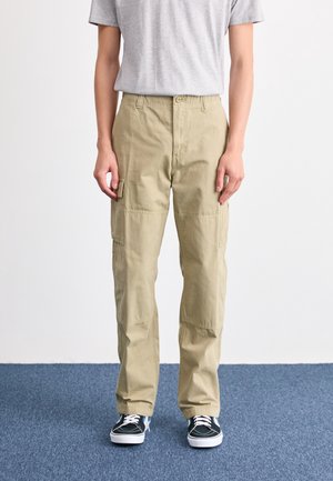 Personne portant un pantalon cargo beige, un t-shirt gris et des baskets noires, debout sur un sol recouvert de moquette bleue contre un mur blanc.