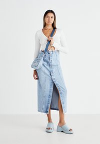 Svetlo modra jeans krilo s sprednjimi gumbi in razporkom do stegna, kombinirano z belo srajco z dolgimi rokavi in svetlo modrimi platformnimi sandali.