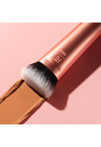 Pinceau de maquillage à tête plate avec des poils noirs et blancs denses appliquant un fond de teint liquide beige sur une surface rose.