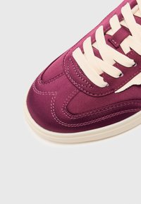 Baskets en satin violet avec lacets crème, dotées d'un design brodé et d'une semelle texturée. Un gros plan montre le matériau lisse et la forme arrondie de la pointe.