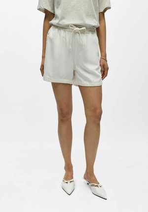 Osoba w luźnych białych szortach ze sznurkiem, jasnoszarej krótkiej koszulce z krótkim rękawem, białych wsuwanych butach na czubku z kokardkami.