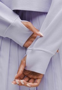 Top de manga larga de color púrpura claro con puños acanalados, mostrado en capas sobre una falda de lavanda con pliegues. Enfoque en el tejido texturizado y los detalles de costura.
