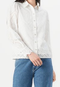 Kvinna i vit ögla spetströja med lång ärm och knappar, tillsammans med blå denimjeans, med handen vilande nära fickan.