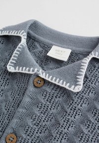 Szürke kötött baba kardigán fehér hímzett gallérszegélyekkel és barna gombokkal, a gallér belsejében "NEXT baby" címkével.