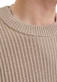 Close-up af en person, der bærer en beige ribstrikket sweaterkrave og et sølvkædehalskæde mod lys hud.