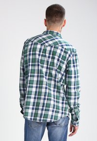 Camisa de algodón a cuadros verdes y azules, diseño de botones, mangas largas, costuras visibles y cuadrados blancos contrastantes en toda la prenda.