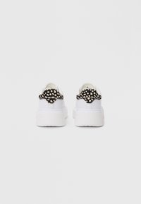 Adidași albi cu o talpă groasă, având un accent texturat cu buline negre și albe pe călcâi. Material neted, design low-top.