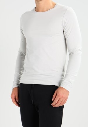 Jasnoszara koszula z długim rękawem wykonana z gładkiego, elastycznego materiału, z okrągłym dekoltem i dopasowanym krojem. Połączona z czarnymi spodniami.