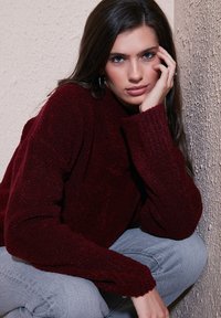 Maroon textured sweater with wide sleeves, ribbed cuffs, paired with light blue denim. Model sits against a textured wall, with hand on chin.