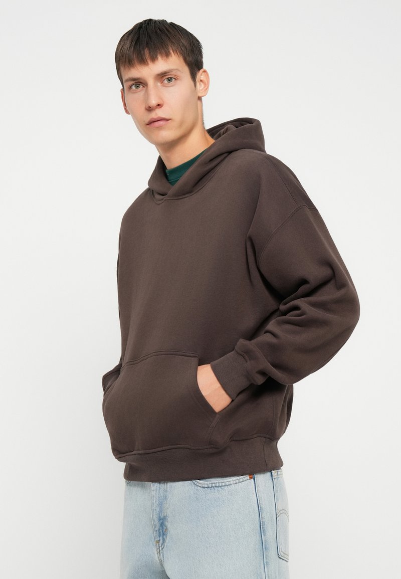 Brauner Kapuzenpullover aus weichem Stoff, mit einer Kängurutasche, lockerer Passform und gerippten Bündchen. Getragen mit hellblauen Jeans.