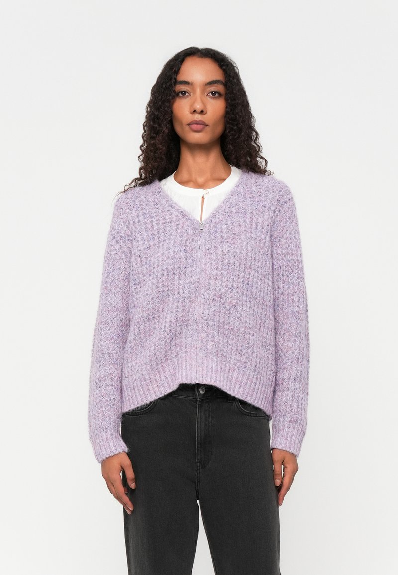 Lavendelfarbener Strickpullover mit einem strukturierten Muster, V-Ausschnitt und geripptem Saum. Kombiniert mit schwarzen Jeans mit hoher Taille, die eine lockere Passform und Taschen haben.