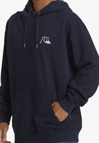 Sudadera con capucha azul marino hecha de tela suave que presenta un pequeño diseño de ola blanca en el pecho. Cuenta con un bolsillo frontal y cordones ajustables.