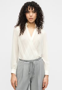 Valge, pika varrukaga pluus V-kaeluse ja rüüstamisdisainiga, tehtud kergest kangast. Sobitatud hallide, voldiliste kõrge vöökohaga pükstega.