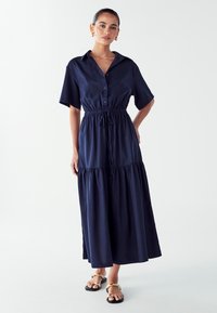 Tamsiai mėlyna, mygtukais užsegama suknelė trumpomis rankovėmis, su susiaurinta juosmens dalimi ir sluoksniuota sijono dalimi. Pagaminta iš lengvos medžiagos su lygiu paviršiumi.