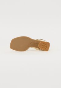 Tan gummisåle med merking; overdelen har et gullfarget stroppdesign. Flat, rund tå med en glatt tekstur og minimal detaljering.