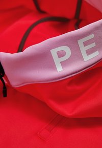 Red sports jacket with a purple collar displaying the word "PE" in large, metallic letters. The fabric appears smooth and stretchy.