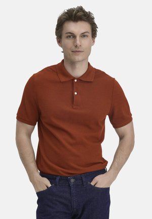 Junger Mann mit heller Haut und braunen Haaren, der ein rostfarbenes Poloshirt und dunkelblaue Jeans trägt und mit den Händen in den Taschen steht.