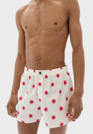 Hombre sin camiseta vestido con calzoncillos blancos con un patrón de rayos de sol rojos sobre un fondo claro, mostrando el torso y las manos.