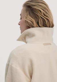 Fleece-Kragenjacke in Off-White mit hohem Kragen und einem Etikett. Weiche Textur mit lockerer Passform und sichtbaren Nahtdetails an den Nähten.