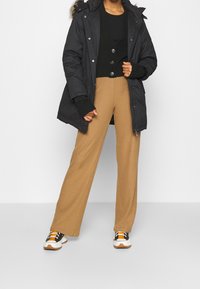 Black parka with a faux fur hood, layered over a black sweater and tan ribbed wide-leg pants, paired with black and white sneakers.