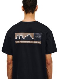 Personne aux cheveux courts et bouclés porte un t-shirt noir avec un motif marron et noir représentant la silhouette d'une ville et le texte « 223 OFF DUTY GEAR F.U.T. B-583 » au dos.