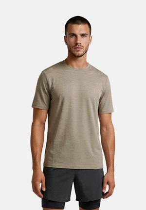 Jonge man met kort haar en baard, die een beige T-shirt met korte mouwen en zwarte korte broek draagt, staand tegen een effen witte achtergrond.