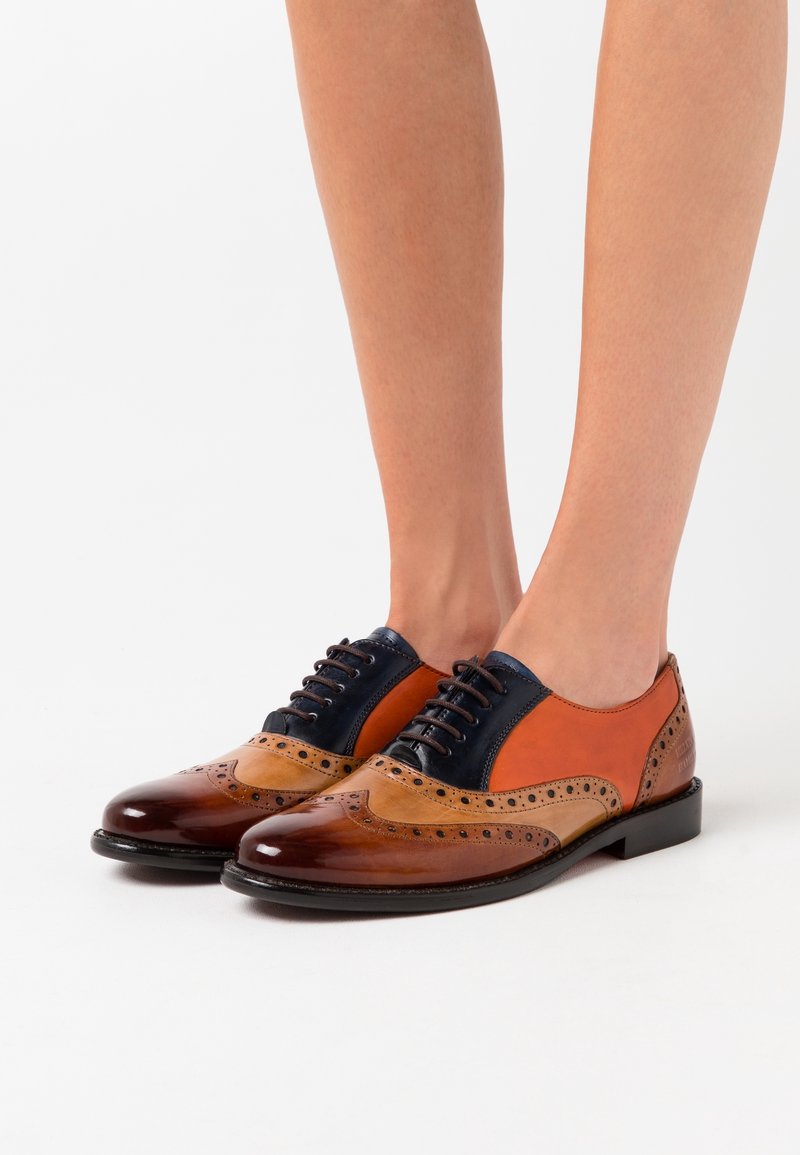 Pieds chaussés de chaussures habillées en cuir brun, beige et bleu marine, de style brogue avec des détails perforés et des lacets noirs sur un fond blanc.