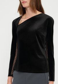 Γυναίκα φοράει μαύρο βελούδινο τοπ με μακριά μανίκια, σχεδιασμό με έναν ώμο και διάφανα μανίκια με πουά δίχτυ, συνδυασμένο με γκρι παντελόνι.