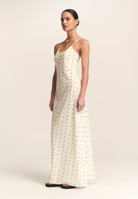Svetlo kremna maksi obleka s tankimi naramnicami, okrašena s črnimi teksturiranimi pikami po celotni površini. Gladek material se elegantno spusti do tal.