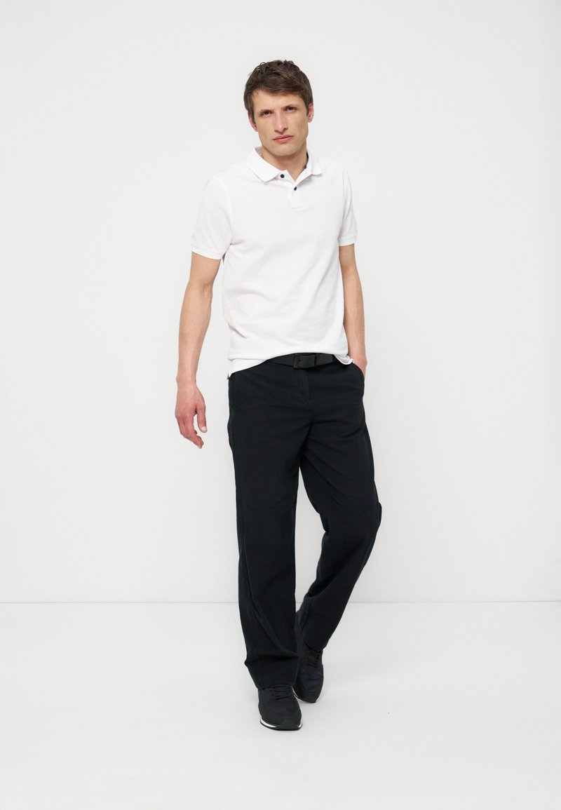 Giovane uomo in piedi con le mani nelle tasche dei pantaloni neri, indossa una polo bianca e sneakers nere su uno sfondo bianco uniforme.