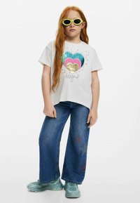 Weißes T-Shirt mit einem mehrfarbigen Paillettenherzdesign und dem Text "Design". Kombiniert mit weiten blauen Jeans mit Sternelementen. Grüne Sonnenbrille.