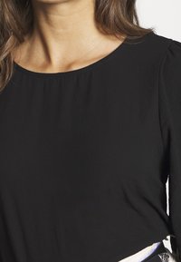 Černý dlouhý rukáv top s kulatým výstřihem, hladkou texturou a nabíranými detaily na ramenních švech. Materiál vypadá lehký a měkký.