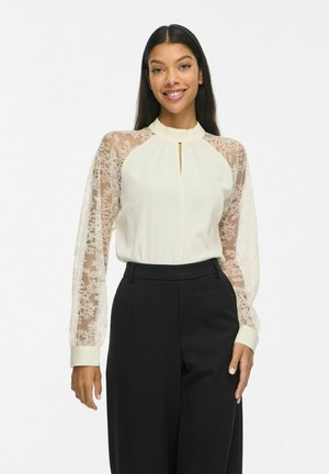 Une femme souriante porte un chemisier couleur crème aux manches en dentelle transparente et un pantalon noir sur un fond blanc uni.