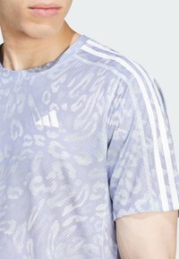 Ljusblå atletisk T-shirt med en texturerad leopardprint, med vita ränder på axlarna och en liten vit logotyp på bröstet.