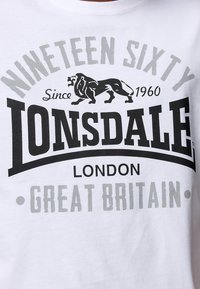 Bijela pamučna majica s velikim crno-sivim grafičkim printom. Tekst uključuje "Lonsdale", "Devetnaest Šezdeset" i ilustraciju lava.