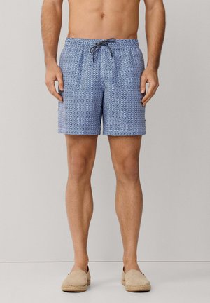 Man die blauwe zwembroek met patroon draagt, voorzien van een zwart trekkoord, en beige instapschoenen, staand tegen een effen achtergrond.