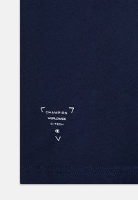 Granatowa bawełniana koszulka z białym nadrukowanym logo, z geometrycznymi akcentami i detalem przeszycia na dolnym brzegu. Prosty, nieozdobiony design.