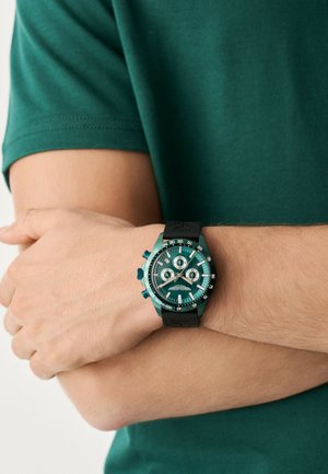 Chronograf hodinky s tyrkysovým ciferníkem, černým gumovým řemínkem, texturovanými akcenty a třemi malými ciferníky. Nosit na zápěstí, s viditelnou zelenou košilí.