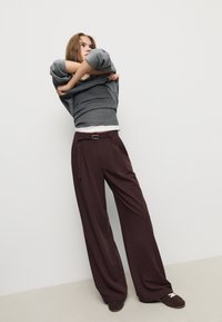 Bruna vida byxor med bälte och sidfickor, kombinerade med en grå långärmad tröja, mot en enfärgad bakgrund.