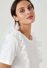 T-shirt bianco con spalle in pizzo arricciato, con motivi floreali e tessuto texturizzato nella parte frontale. Semplice scollatura rotonda.