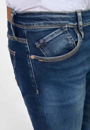 Jeans in denim di colore blu scuro con cuciture visibili, cinque tasche e una piccola etichetta con logo sulla tasca anteriore. Tessuto liscio, design aderente.