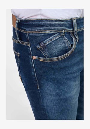 Vaqueros de mezclilla en azul oscuro con costuras visibles, cinco bolsillos y una pequeña etiqueta de logo en el bolsillo delantero. Textura suave, diseño ajustado.