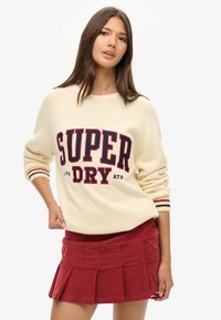Strikcreme sweater med "SUPER DRY" logo i rød og navy, ribbet manchetter. Kombineret med en rød fløjl plisseret miniskirt.